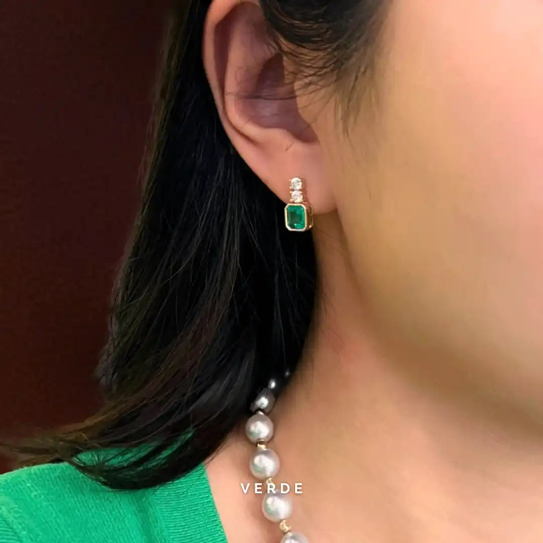 Close-up of an earring with a green gemstone and pearl drop, worn by a person with dark hair.